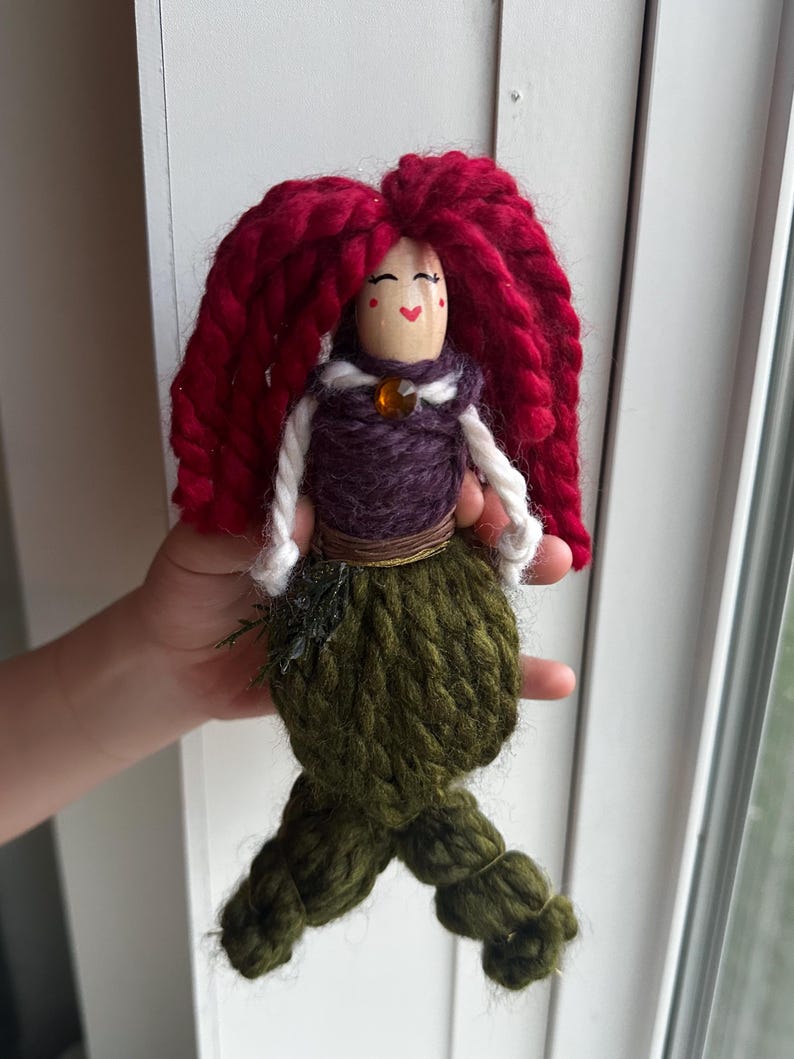 May include: A handmade yarn doll with red hair, a purple bodice, and a green mermaid tail. The doll has a painted face with red cheeks and a smile.