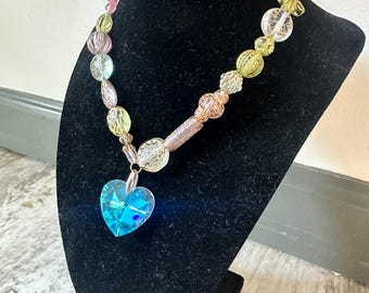 Puede incluir: Un collar de cuentas con un colgante de cristal azul en forma de corazón. El collar presenta una variedad de cuentas de colores en tonos rosa, verde y transparente. El collar se exhibe en un busto de terciopelo negro.