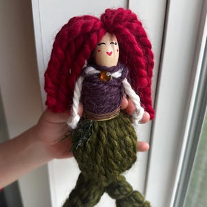 May include: A handmade yarn doll with red yarn hair, a purple yarn bodice, a green yarn tail, and a wooden head with a painted face.