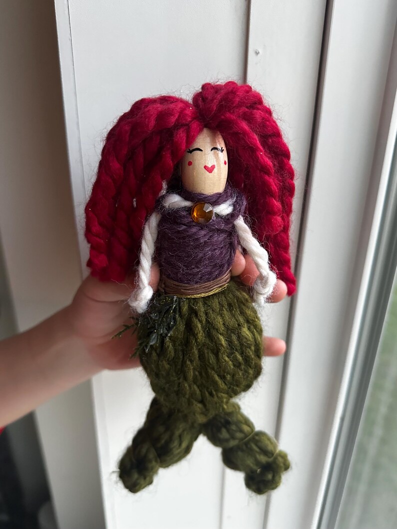 May include: A handmade yarn doll with red yarn hair, a purple yarn bodice, a green yarn tail, and a wooden head with a painted face.