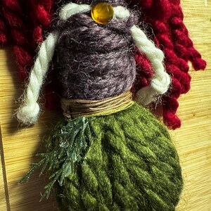 May include: A handmade yarn doll with red hair, a purple bodice, a brown belt, and a green knitted tail. The doll has a wooden head with painted eyes and a heart on the forehead. The doll is wearing a white yarn necklace with an amber bead.