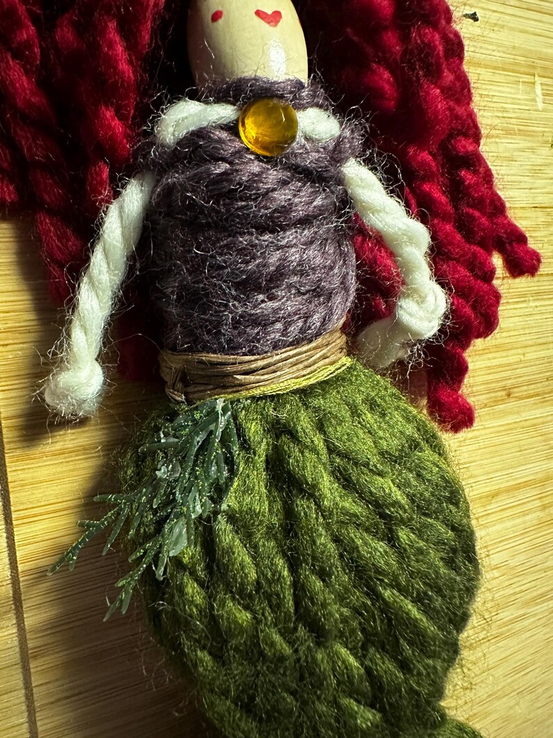 May include: A handmade yarn doll with red hair, a purple bodice, a brown belt, and a green knitted tail. The doll has a wooden head with painted eyes and a heart on the forehead. The doll is wearing a white yarn necklace with an amber bead.