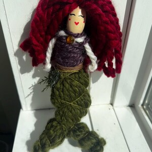 May include: A handmade yarn doll with red hair, a purple and white torso, and a green yarn tail. The doll has a painted face with red cheeks and a small orange bead for a necklace.