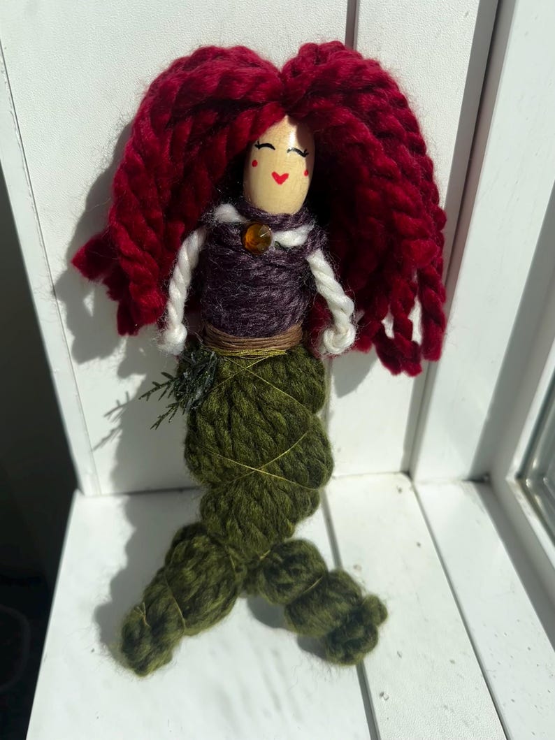 May include: A handmade yarn doll with red hair, a purple and white torso, and a green yarn tail. The doll has a painted face with red cheeks and a small orange bead for a necklace.