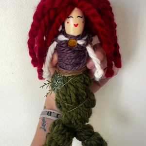 May include: A handmade doll with red yarn hair, a purple yarn bodice, and a green yarn tail. The doll has a painted face with red cheeks and a smile.