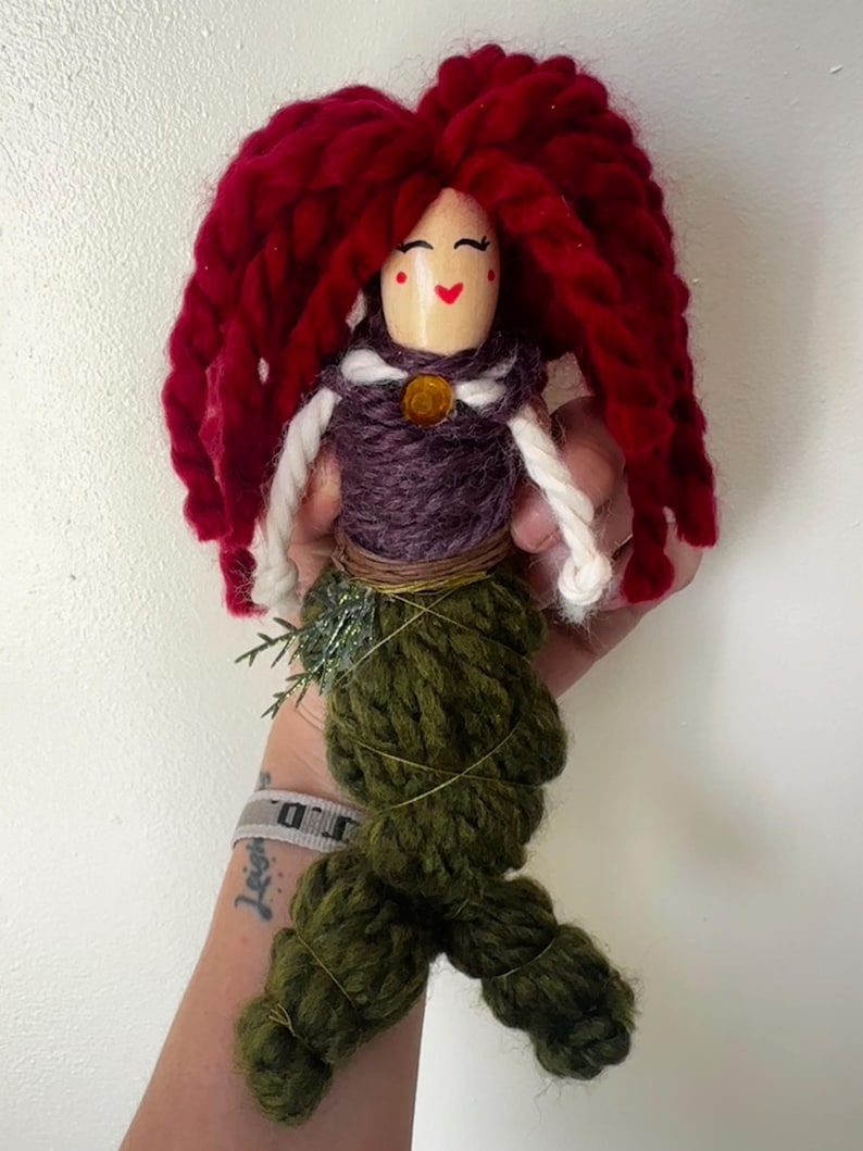May include: A handmade doll with red yarn hair, a purple yarn bodice, and a green yarn tail. The doll has a painted face with red cheeks and a smile.
