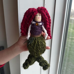 May include: A handmade yarn doll with red yarn hair, a purple yarn bodice, and a green yarn tail. The doll has a painted face with red cheeks and a small orange bead for a nose.