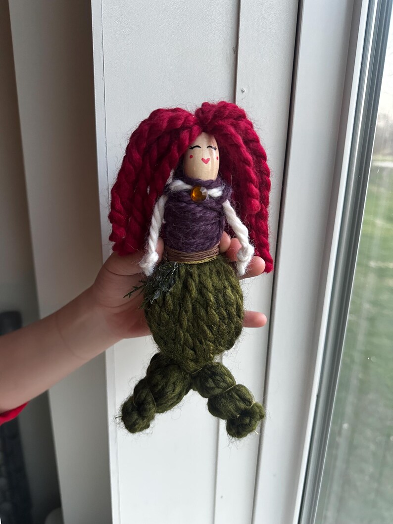 May include: A handmade yarn doll with red yarn hair, a purple yarn bodice, and a green yarn tail. The doll has a painted face with red cheeks and a small orange bead for a nose.