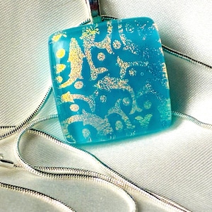 May include: A square, turquoise pendant with gold-colored, abstract patterns hangs on a silver chain. The pendant is made of glass and has a shiny, reflective surface. The necklace is displayed on a white, textured background.