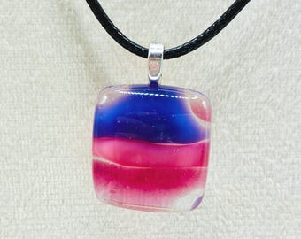 May include: A square pendant necklace with a layered design of blue, pink, and clear glass. The pendant hangs from a black cord with a silver-coloured bail. The glass has a glossy finish.