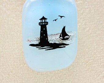 May include: A light blue glass bottle with a coastal design. The design features a black silhouette of a lighthouse, a sailboat, and three birds. The bottle has a gradient effect, transitioning from a darker blue at the top to a lighter blue at the bottom.