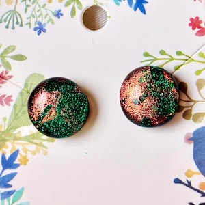 May include: A pair of round stud earrings featuring a dichroic glass surface with a mottled pattern of metallic copper and vibrant emerald green. The earrings are mounted on a white display card decorated with a colorful floral and bird print.