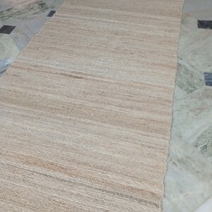 May include: A natural fiber rug with a woven texture in a light beige color. The rug is laid out on a tiled floor.