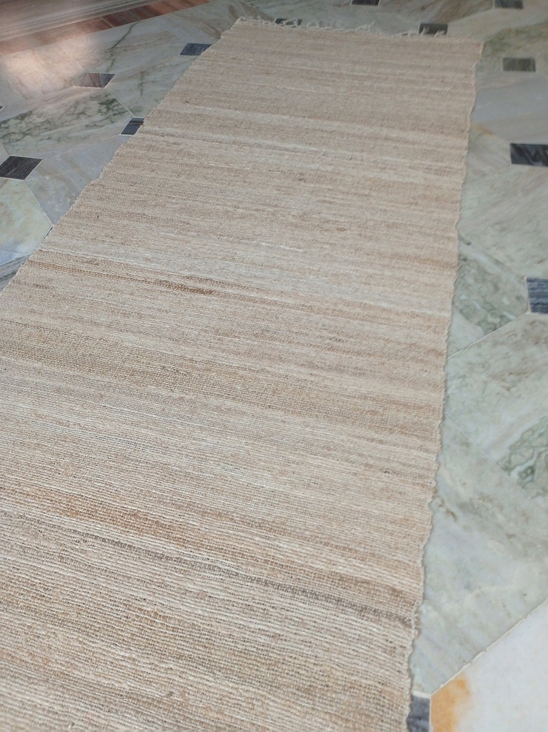 May include: A natural fiber rug with a woven texture in a light beige color. The rug is laid out on a tiled floor.