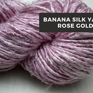 Puede incluir: Un ovillo de hilo de seda de pl&aacute;tano de color oro rosa. El hilo es suave y delicado, con un brillo sutil.