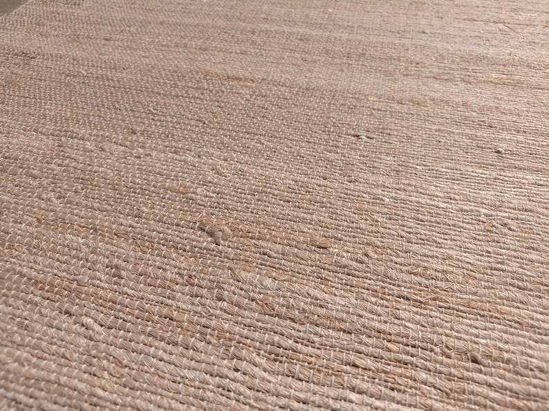 May include: Close-up of a natural fiber rug with a woven texture. The rug is a light brown color and has a subtle sheen.