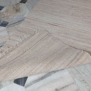 May include: A beige woven rug with a fringed edge. The rug is folded in half and laid on a tiled floor.
