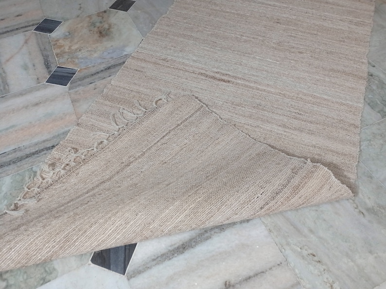 May include: A beige woven rug with a fringed edge. The rug is folded in half and laid on a tiled floor.