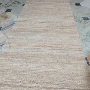 May include: A natural fiber rug with a woven texture in a light beige color. The rug has a frayed edge and is laid out on a tiled floor.