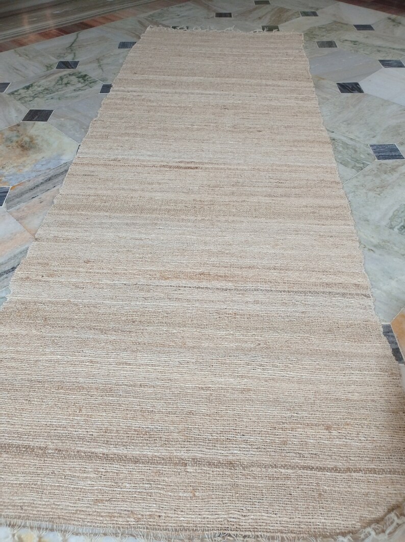 May include: A natural fiber rug with a woven texture in a light beige color. The rug has a frayed edge and is laid out on a tiled floor.