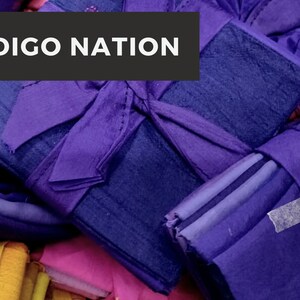 Op de afbeelding: Een stapel indigo-gekleurde vierkante stoffen, sommige gebonden met een paars lint, met de tekst "INDIGO NATION" bovenaan de afbeelding.