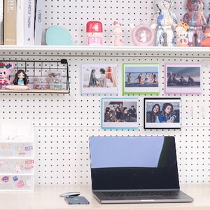 May include: A white pegboard with a variety of framed photos, including a group of people, a couple, and a beach scene. There is a laptop computer on a white desk in front of the pegboard.