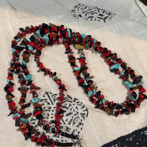 May include: A multi-strand necklace composed of small, irregularly shaped beads in red, black, turquoise, and brown. The necklace is displayed on a cream-colored fabric with a black and white pattern.
