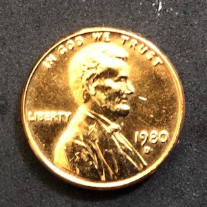 May include: A gold-colored 1980 US penny coin with the words "In God We Trust" and "Liberty" inscribed around the edge. The coin features a profile of Abraham Lincoln.