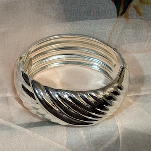 May include: A silver bangle bracelet with a ribbed design. The bracelet features a hinged clasp and a curved, textured section. The jewellery is displayed on a light-coloured, patterned fabric.