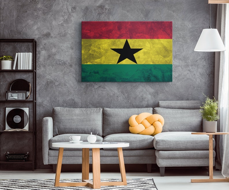 May include: A gray couch with a yellow knotted pillow in front of a gray wall with a distressed canvas print of the flag of Ghana. The flag is red, yellow, and green with a black star in the center.