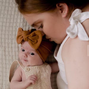 May include: A woman gently kisses a baby wearing a brown turban with a bow on top and a cream knitted onesie.