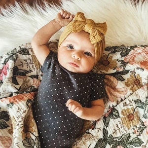 May include: A baby wearing a black and white polka dot onesie and a mustard yellow knotted headband. The baby is lying on a white and floral patterned blanket.