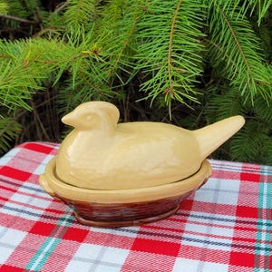 May include: A ceramic duck-shaped dish with a lid, in shades of tan and brown. The duck sits in a nest-like base, placed on a red and white checkered tablecloth. The background features green pine branches.