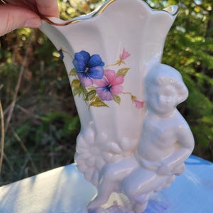 May include: A white porcelain vase with a gold rim, featuring a floral design of blue and pink pansies. A small cherub figure is sculpted on the side. The vase is held by a hand, with a blurred natural background.