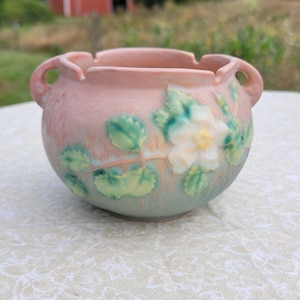 May include: A pink and green glazed ceramic planter with a raised design of a white rose and leaves. The planter has two handles on the sides.