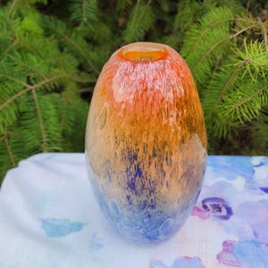 May include: An oval-shaped glass vase with a gradient of colors. The top is orange, transitioning to brown, and then to blue at the base. The vase has a textured surface and is likely used for decorative purposes.