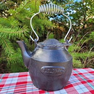 May include: A black cast iron kettle with a silver wire handle. The kettle has a spout and a lid. The brand name "L.L.Bean" is embossed on the side. The kettle is sitting on a red and white checkered tablecloth.