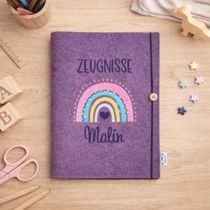 Könnte beinhalten: Ein lila Filz-Notizbuchcover mit dem Wort "ZEUGNISSE" in Blau gestickt. Unter dem Text befindet sich ein Regenbogen-Design mit einem Herz, und der Name "Malin" ist unter dem Regenbogen gestickt. Ein schwarzes Gummiband mit einem Holzknopfverschluss sichert das Notizbuch.