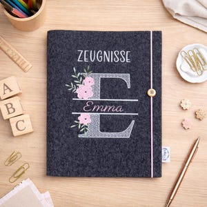 Könnte beinhalten: Eine dunkelgraue Filzhülle mit dem Wort "ZEUGNISSE" in Weiß gestickt. Der Buchstabe "E" ist mit rosa Blumen verziert und der Name "Emma" ist darunter gestickt. Ein rosa Band und ein Holzknopf sichern die Hülle.