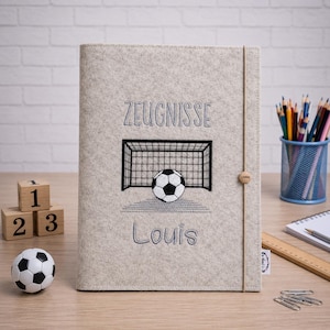 Könnte beinhalten: Eine hellgraue Dokumentenmappe aus Filz mit einer Stickerei eines schwarz-weißen Fußballtors und Balls. Über dem Tor ist der Schriftzug "ZEUGNISSE" und darunter der Name "Louis" eingestickt. Ein dünnes Gummiband hält die Mappe sicher verschlossen.