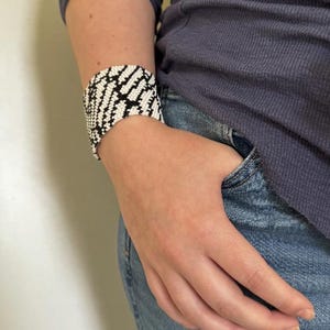 May include: A wide cuff bracelet with a geometric pattern in black and white. The bracelet is made of small beads and is worn on the wrist. The person is wearing a blue long-sleeve shirt and blue jeans.