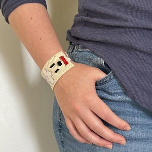 May include: Cream-colored beaded cuff bracelet with a whimsical design. The bracelet features black and red accents, creating a playful contrast. This accessory adds a unique touch to any outfit.