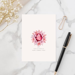 Può includere: Biglietto di auguri con un'illustrazione ad acquerello di un rubino circondato da fiori rosa. La carta include il testo "July, Ruby & Water Lily, Passion - Confidence - Vitality". Sono presenti anche una penna nera e una graffetta dorata.
