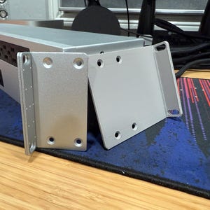 May include: Three silver metal brackets with rounded corners and multiple screw holes. The brackets are on a blue and red patterned surface.