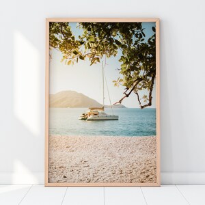 May include: A framed photograph of a white sailboat anchored in a turquoise bay with a sandy beach in the foreground. Lush green trees hang over the water in the background.