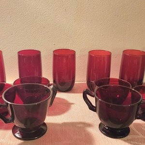 May include: Set of eight ruby red glass cups, four are tall and straight, and four are shorter with two handles. The cups are arranged on a table with a floral pattern.