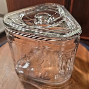 May include: A clear glass jar with a triangular lid. The jar has a rectangular body with etched details and a circular indentation on the lid. The jar is sitting on a wooden surface.