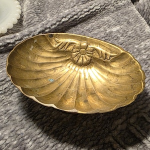 May include: A vintage brass seashell-shaped dish with a floral design in the center. The dish has a scalloped edge and a golden hue, showing signs of age and use. The dish is likely used for holding small items.