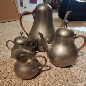 Może przedstawiać: Zestaw do herbaty z cyny w stylu vintage, w tym czajniczek, dzbanek do kawy, śmietanka i cukiernica. Czajniczek i dzbanek do kawy mają plecione uchwyty. Zestaw jest prezentowany na teksturowanej, jasnobrązowej powierzchni.