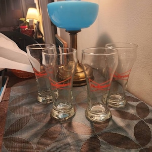 May include: Four clear glass beer mugs with the word "Budweiser" printed in red on the front. The mugs are sitting on a brown and blue patterned placemat.